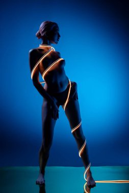 Bir kadın zarif bir şekilde üstsüz poz verir, vücudu parlak mavi bir zemin karşısında parıldayan ışığa sarılır, büyüleyici bir görsel karşıtlık yaratır..