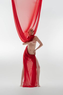 Bir kadın güzelliğini minimalist bir ortamda sergiliyor, kırmızı kumaşa bürünmüş, üstsüz poz verirken kendine güven ve zarafet saçıyor..