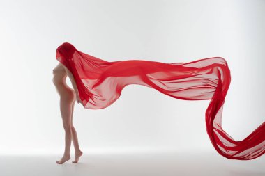 Bir kadın zarif bir şekilde üstsüz duruyor. Havada zarif bir şekilde ilerleyen kırmızı kumaşla süslenmiş. Yumuşak, aydınlık bir arkaplan üzerine kurulmuş..