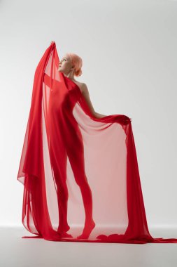 Bir kadın şeffaf kırmızı kumaş içinde zarif bir şekilde duruyor, güzelliği ve sadeliği minimalist bir ortamda harmanlıyor..