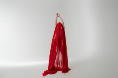 Bir kadın zarif bir şekilde poz verir. Kollarını kaldırır ve saf kırmızı bir kumaşa sarınır. Tarafsız bir arkaplanda güzelliği ve zarafeti ön plana çıkarır..