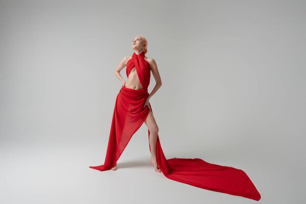 Женщина уверенно стоит, украшенная струящимися красными тканями, которые подчеркивают ее красоту и грацию в хорошо освещенной студии.
