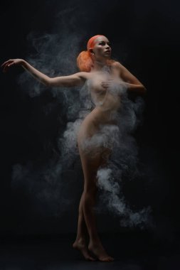 Kendine güvenen kızıl saçlı bir kadın çıplak duruyor, kısmen kıvrımlı kumaş ve dumanla gizlenmiş, güzelliğini sergiliyor..