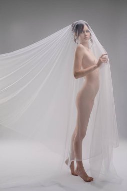 Bir kadın zarif bir şekilde duvak takmış, minimalist bir ortamda güzelliğini ve özgüvenini ön plana çıkarıyor, şehvet ve cazibeyi çağrıştırıyor..