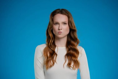 Bir kadın stüdyoda canlı bir mavi arkaplana karşı duruyor, düşünceli bir ifade sergiliyor. Uzun, dalgalı saçları, iç gözlem anını yakaladığında yüzünü çerçeveler..