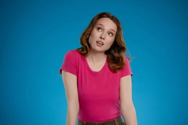 Bir stüdyo ortamında, bir kadın neşeli bir ifadeyle poz verir, uzun saçları yavaşça omuzlarına düşer. O parlak pembe bir gömlek giyer, mavi bir arka planla çelişir..