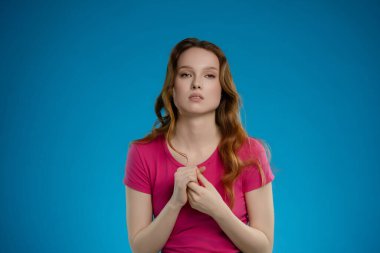 Bir kadın stüdyoda duruyor, yansıtıcı bir duygu sergiliyor. Uzun saçları, elleri özenle sıkıca sarılmış, canlı mavi bir arkaplan tarafından çerçevelenmiş bir şekilde poz verirken nazikçe akıyor..