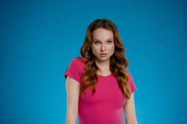 Bir kadın kendine güvenerek stüdyoda duruyor, pembe bir bluz giyiyor ve saçları omuzlarının üzerinden dökülüyor. İfadesi bir duygu karışımı taşıyor, canlı mavi bir arkaplan karşısında dikkatleri çekiyor..