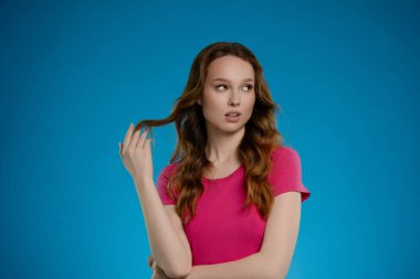 Bir stüdyo ortamında, dalgalı saçlı bir kadın açık mavi bir arkaplana karşı şakacı bir şekilde poz verir, kilitlerini okşarken merak ve güven karışımı gösterir..