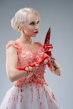 Bir stüdyo çekimi sırasında eşsiz gelinlik giymiş bir kadın model. Elbise karmaşık detaylarla süslenirken, elleri ve bıçağı kanla lekelenmiş, yumuşak arka planda dramatik ve ürkütücü bir kontrast oluşturuyor..