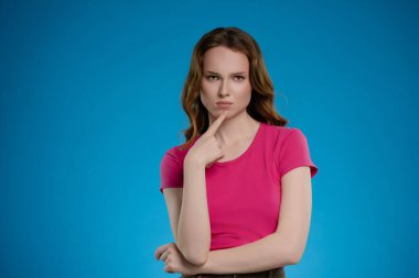 Bir kadın stüdyoda canlı mavi bir arka planla duruyor, eli çenesinde düşünceli bir şekilde poz veriyor. İfadesi, derin düşüncelere dalma ve duygu karışımı olarak izleyiciyi kendine çekiyor..