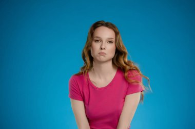 Dalgalı saçı ve pembe bluzuyla stüdyo ortamında dikkat çeken bir kadın. Düşünceli bir şekilde poz veriyor, çarpıcı bir mavi zemine karşı bir duygu duygusu yaratıyor..