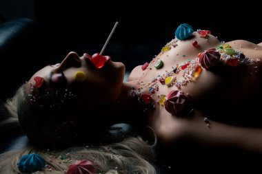 Bir kadın karanlık bir yüzeyde uzanır. Şeker temalı vücut sanatı ve ışığı yakalayan canlı dekorasyonlarla süslenir..