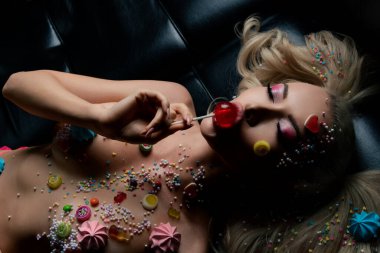 Tatlı temalı vücut sanatı ile süslenmiş bir kadın lolipopun tadını çıkarır, şekerlerle kaplı bir arka planda uzanır..