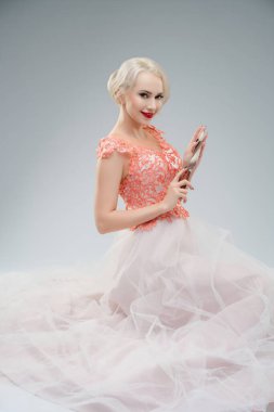 Göz alıcı bir gelinlik giymiş, üzerinde kan lekeleri olan bir kadın profesyonel bir stüdyo ortamında güven içinde poz veriyor. İfadesi büyüleyici ve ilgi çekici. Gelinliğinin narin özellikleriyle çelişiyor..