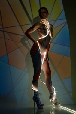 Bir güzellik, vücuduna karşı renkli desenler gösteren parlak projektör ışığıyla aydınlatılmış üstsüz duruyor..