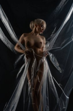 Stüdyoda bir kadın dikiliyor. Saf bir kumaşın içinde zarifçe poz veriyor. Yumuşak kumaş vücudunu sarıp ışıkta narin şekiller ve yansımalar oluşturuyor. Koyu arkaplan, sahnenin çarpıcı görüntüsünü güçlendirir..