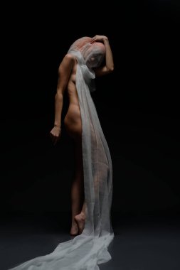 Bir kadın zarif bir şekilde çıplak poz verir, akıcı kumaşla örtülmüş, koyu arkaplan ile çarpıcı bir kontrast yaratır..