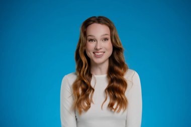 Bir kadın stüdyoda canlı bir mavi arkaplana karşı duruyor, gülümsemesiyle neşeyi ifade ediyor. Saçları dalgalar halinde dalgalanıyor, sıcak bir şekilde, kendine güven ve duygu yayıyor..