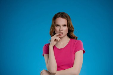 Bir stüdyo ortamında, bir kadın parlak mavi bir arka plana karşı poz verir, parmağını hafifçe ısırırken düşünceli bir yüz ifadesi sergiler. Sıradan pembe bir bluz giyiyor..