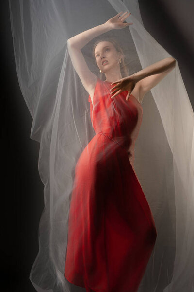 A woman is posing gracefully in a studio, wrapped in soft drapery that enhances her elegant red dress. Her expression conveys deep emotion amidst the flowing fabric.