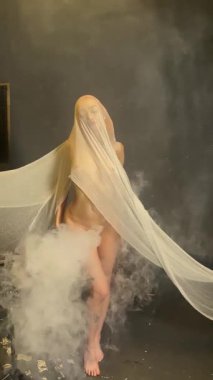 Göz kamaştırıcı bir kadın şeffaf kumaşla etkileşime girer. Sisli, şık bir stüdyoda büyüleyici bir görüntü yaratır..