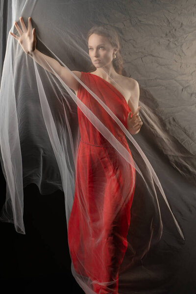 In a studio setting, a woman elegantly poses in a striking red dress, using sheer fabric to accentuate her graceful movements. Her expression conveys deep emotion, inviting viewers to connect.