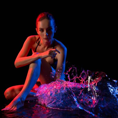 Parlak su ile oturan bir model sergiler dramatik kırmızı ve mavi ışıklandırma efektleri, pozunu ve su akışını vurgular.