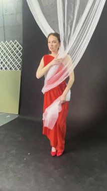 Bir kadın zarif bir şekilde stüdyoda poz verir, kırmızı bir elbise ve topuklu ayakkabı giyer..