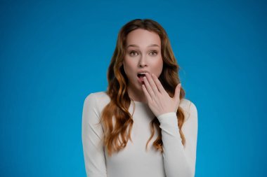 Bir kadın stüdyoda durmuş, şaşırmış bir ifade sergiliyor. Uzun, dalgalı saçları var ve elini ağzına kaldırıyor. Parlak mavi bir zemin karşısında şaşkınlık hissi veriyor..