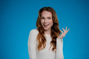 İyi ışıklandırılmış bir stüdyoda, uzun, dalgalı saçlı bir kadın neşeli bir yüz ifadesi sergilerken, açık mavi bir arkaplanda şakayla poz veriyor. Hareketleri sıcak bir etkileşim olduğunu gösteriyor..