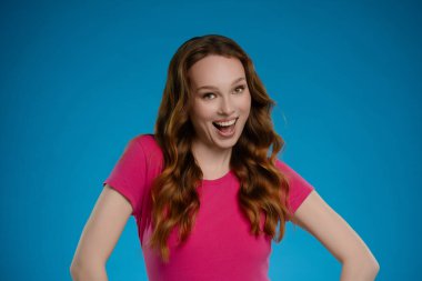 Bir kadın kendine güvenerek stüdyoda duruyor, parlak pembe bir gömlek giyiyor ve neşeli bir ifade sergiliyor..