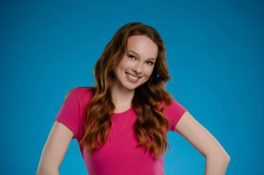 Bir kadın, pembe bir bluz giyerken, parlak bir gülümseme sergileyerek, kendine güvenerek stüdyoda duruyor. Dalgalı saçları canlı mavi arkaplanı tamamlıyor ve canlı bir atmosfer yaratıyor..