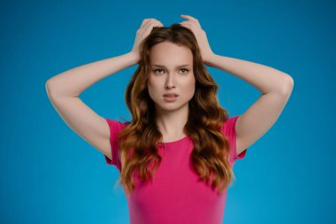 Bir kadın stüdyoda poz verirken görülüyor, elleri saçlarında çeşitli duygular ifade ediyor. Parlak mavi arka plan onun duygusal ifadesini arttırıyor..