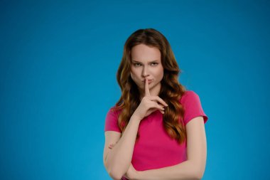 Bir kadın kendine güvenerek stüdyoda duruyor, pembe bir gömlek giyiyor ve duygularını dudaklarına yansıtıyor, sessizliği işaret ediyor. Canlı mavi arkaplan çarpıcı pozu güçlendirir.