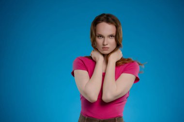 Bir kadın stüdyo ortamında güçlü bir ifade sergiliyor. Ellerini boynuna dolayarak duygusal derinliğini çarpıcı mavi arka plana vuruyor..