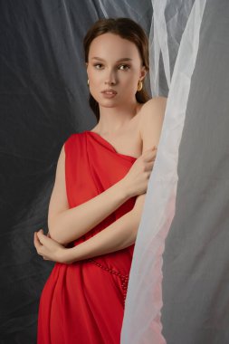 Akan kırmızı elbiseli bir kadın stüdyoda sakin bir ifadeyle poz veriyor. Saf bir arkaplanın yanında duruyor, büyüleyici bir atmosfer yaratıyor ve zarafetini sergiliyor..