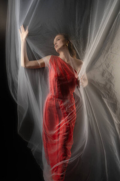 A woman poses against a backdrop while interacting with flowing fabric. Her expression captures deep emotion, enhanced by the studio lighting that creates a dreamlike atmosphere.