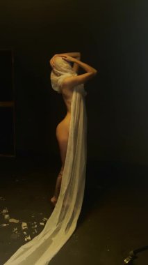 Kırmızı elbiseli bir kadın loş bir stüdyoda yumuşak kumaşla poz veriyor, güzellik ve sanatın altını çiziyor..