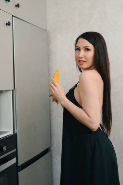 Siyah elbiseli, uzun siyah saçlı bir kadın modern bir mutfakta elinde mango tutuyor. Kadın kameradan uzağa bakıyor, omzunun üzerinden bakıyor. Mutfak sade ve şık, paslanmaz çelik buzdolabı ve siyah fırını var..