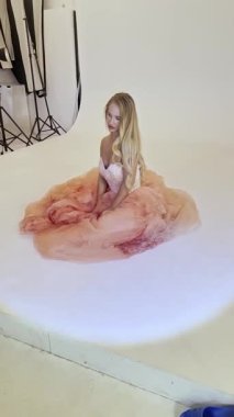 Zarif bir kadın stüdyoda oturuyor. Etrafı dalgalı kumaşlarla çevrili. Duruşunda güzellikler ve sanatsal yetenekler sergiliyor..