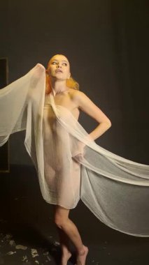 Canlı kızıl saçlı bir kadın stüdyoda saf kumaşla zarif bir şekilde etkileşime geçip güzelliğini ve yaratıcılığını ön plana çıkarıyor..