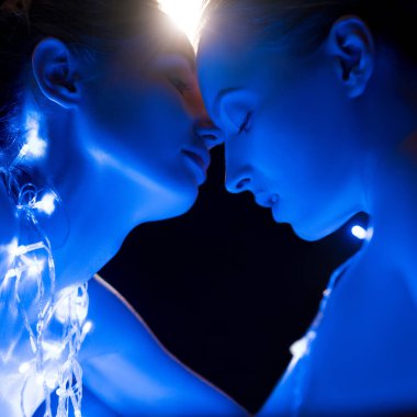 Yumuşak mavi ışıklar, iki bireyin birbirine yakın eğilmesiyle huzurlu bir atmosfer yaratır. İfadeleri, bağlantı ve yakınlığı ifade eder..