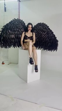 Bir kadın özgüvenli bir şekilde stüdyoda poz verir, özenle hazırlanmış siyah kanatlar ve yaratıcı bir ortamda şık kıyafetler sergiler..