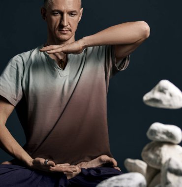 Yogada oturan bir adam stüdyonun yanında beyaz taşlarla poz veriyor.