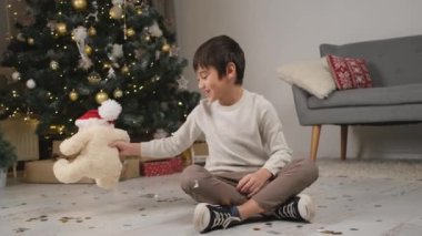 Küçük çocuk Noel süsü verilmiş odada oyuncak ayıyla oynuyor.