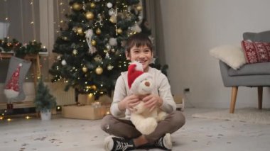 Küçük çocuk Noel süsü verilmiş odada oyuncak ayıyla oynuyor.