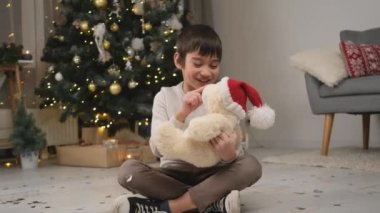 Mutlu küçük çocuk Noel ağacıyla odada oyuncak ayıyla oynuyor.