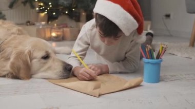 Çocuk Noel Baba 'ya mektup yazarken Golden Retriever köpeğinin yanında Noel süsü verilmiş bir odada yatıyordu.
