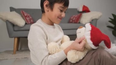 Küçük çocuk Noel kutlaması için dekore edilmiş odada oyuncak ayıya örülmüş Noel Baba şapkası takıyor.